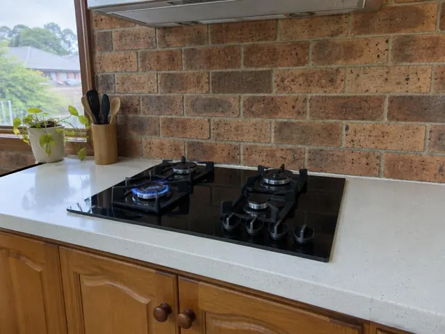 Black gas cooktop set into a light benchtop with brick splashback and wood cabinets in Tingira Heights
